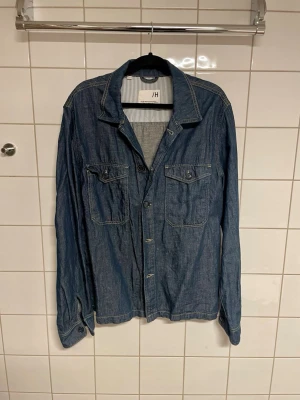 Blå jeansjacka från select homme - Säljer en klassisk blå jeansjacka från select home med två bröstfickor med knappar och snygga sömdetaljer. Jackan har krage, knäppning framtill och långa ärmar. Perfekt för dig som gillar en tidlös och avslappnad stil.