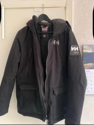 Svart Helly Hansen jacka - Cool svart jacka från Helly Hansen med logga på bröstet och ärmen. Jackan har huva, dragkedja och två stora fickor framtill. Perfekt för att hålla vinden borta och ge en sportig look. Materialet är slitstarkt och jackan har en modern passform.