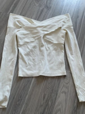 Offshoulder vit  topp - Trendig offshoulder topp i vit ribbad bomullsmix. Långärmad och figurnära passform med snygg omlottkänsla över bysten. Perfekt för dig som vill ha en stilren och modern look.