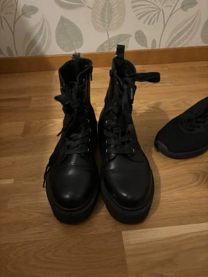 Svarta chunky kängor med snörning - Säljer ett par svarta kängor med chunky sula och snörning framtill. Kängorna har dragkedja på insidan och är tillverkade i ett glansigt skinnliknande material. Perfekta för dig som vill ha en edgy och trendig look. Använda en sommar men för små nu. Fortfarande i jättefint skick. 