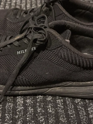 Svarta Tommy Hilfiger sneakers - Snygga helsvarta sneakers från Tommy Hilfiger med stickad ovandel och diskret logga på sidan. Skorna har svarta snören, en bekväm sula och en liten detalj med Hilfigers färger bak på hälen. Perfekta för en clean och stilren look.