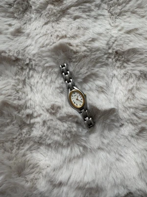 Silverfärgad Seiko damklocka - Säljer en elegant damklocka från Seiko med silverfärgat armband och oval urtavla. Urtavlan har romerska siffror och en subtil gulddetalj runt kanten. Perfekt för dig som gillar klassisk och stilren design. 