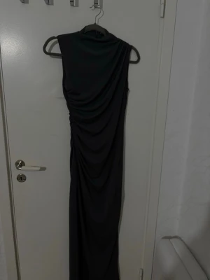 Mörkgrå långklänning med drapering - Säljer en grå lång klänning med draperad detalj på sidan. Klänningen är ärmlös och har en elegant, figurnära passform. Materialet är mjukt och stretchigt, vilket ger en snygg siluett. Perfekt för dig som gillar stilrena och trendiga plagg.