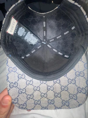 Gucci keps med GG-mönster - Säljer en snygg Gucci keps i beige med blått GG-monogram över hela kepsen. Klassisk böjd skärm och ikoniskt rött och blått band på sidan. Tillverkad i canvasmaterial med detaljer i svart tyg på insidan. Perfekt för dig som vill ha en exklusiv och trendig look.