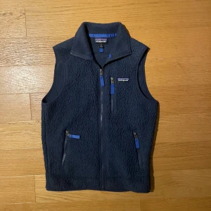 Mörkblå fleeceväst från Patagonia XS - Säljer en mörkblå fleeceväst från Patagonia i storlek XS. Västen har hög krage, tre fickor med dragkedja och ett mjukt, fluffigt material. Perfekt för lager-på-lager och outdoor vibes. Diskret Patagonia-logga på bröstet och insidan.