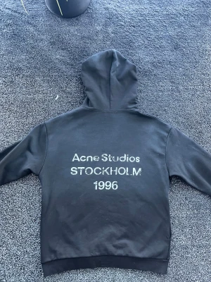 Acne studios hoodie  - Svart hoodie från Acne Studios med tryck på ryggen där det står 'Acne Studios STOCKHOLM 1996'. Klassisk passform med huva och ribbade muddar. Perfekt för dig som gillar stilrena plagg med en touch av streetwear. Materialet känns mjukt och skönt. Storlek M, skick 9/10. Pris går att diskutera✅