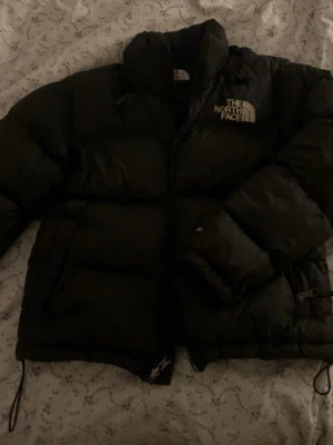 Svart pufferjacka från The North Face - Säljer en svart pufferjacka från The North Face med logga på bröstet och ryggen. Jackan har hög krage, dragkedja och justerbara snören i nederkant. Perfekt för kalla vinterdagar och riktigt trendig stil. Fast den har ett litet hål i den innersta fickan som går att sy 