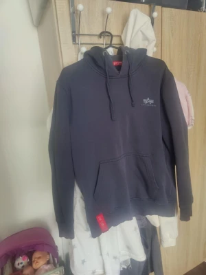 Mörkblå hoodie från Alpha Industries - Säljer en mörkblå hoodie från Alpha Industries med klassisk logga på bröstet och röd tag på fickan. Tröjan har en stor känguruficka, ribbade muddar och justerbar huva med snören. Perfekt för dig som gillar streetwear och enkel stil.