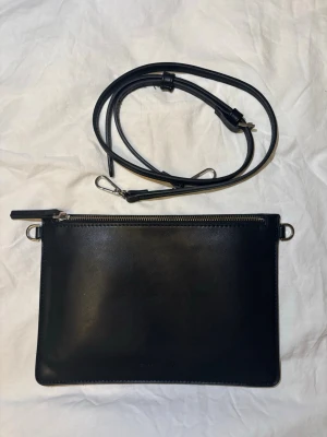 Svart kuvertväska från Carin Wester - Populär svart axelväska från Carin Wester i slätt skinn. Väskan är i bra skick och har används ett fåtal gånger. Väskan har en avtagbar och justerbar axelrem, silverfärgade metalldetaljer och dragkedja upptill. Insidan har ett fack med dragkedja och plats för kort. Perfekt för dig som gillar stilren design.                                                                                     Priset kan diskuteras!