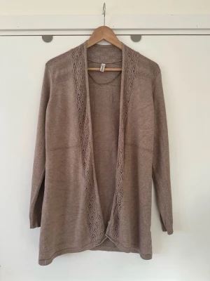 Brun Soyaconcept kofta, storlek S / M - Mysig beige kofta med öppen front och fina spetsdetaljer längs kanterna och axlarna. Långärmad och i mjukt stickat material, perfekt för lager-på-lager. Enkel och stilren design som passar till många outfits. Soyaconcept, storlek S / M. 