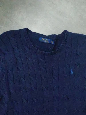 Ralph Lauren kabelstickad - Kabelstickad tröja i mörkblått från Polo Ralph Lauren med rund halsringning och den klassiska broderade loggan på bröstet. Tröjan har ribbade muddar och är tillverkad i mjukt material som passar perfekt till en avslappnad men stilren look.