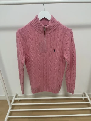 Rosa kabelstickad tröja Polo Ralph Lauren - Säljer en rosa kabelstickad tröja från Polo Ralph Lauren med halv dragkedja och hög krage. Tröjan har det klassiska broderade logotypen på bröstet och ribbade muddar vid ärmslut och nederkant. Perfekt för dig som gillar preppy stil.