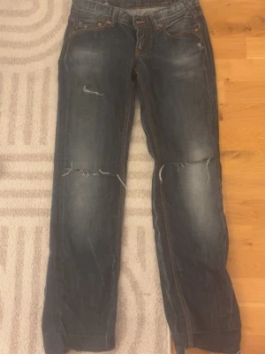 Mörkblå slitna jeans med raka ben - Säljer ett par mörkblå jeans med raka ben och slitningar på både lår och knän. Jeansen har klassisk femficksdesign, kontrastsömmar och en avslappnad passform. Perfekta för en cool och avslappnad stil.