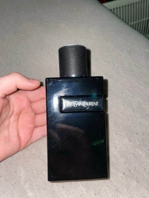 Yves Saint Laurent svart parfymflaska - Snygg parfymflaska från Yves Saint Laurent i svart glas med stilren, rektangulär form och metallisk kork. Märkets logga syns tydligt på flaskan och korken har en graverad YSL-logga på toppen. Perfekt för dig som gillar exklusiva dofter.