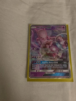 Pokémonkort Mewtwo GX - Säljer ett Pokémonkort med Mewtwo GX. Kortet har en färgstark design i lila och rosa toner, med holografiska effekter och tydliga attacker som Telekinesis, Reigning Pulse och Psychic Nova GX. Kortet är inplastat i ett gult skyddsfodral.