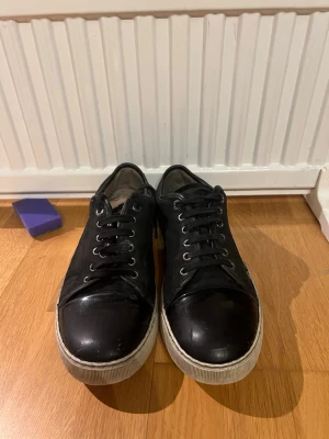 Svarta Lanvin sneakers i läder - Snygga svarta sneakers från Lanvin med ovandel i läder och tåparti i blank finish. Skorna har vita sulor med Lanvin-logga undertill och svarta skosnören. Klassisk låg modell som passar till många olika stilar.