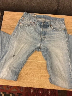 Levi's 501 ljusblå  - Ljusblå levis 501 med slitningar och hål på höger lår. De har knappgylf. Vid minsta fundering är det bara att skriva.