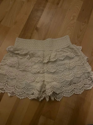 Vita virkade shorts med volanger - Säljer ett par vita virkade shorts med bred resår i midjan och flera lager volangdetaljer. Shortsen har en romantisk och bohemisk känsla och passar perfekt till sommaren. Superfina att matcha med en enkel topp eller crop top.