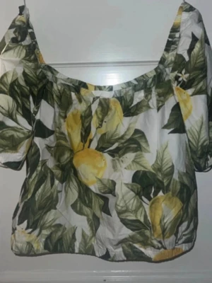 Blommig offshoulder topp med citroner - Säljer en söt offshoulder topp med citronmönster i gult och grönt på vit botten. Toppen har korta puffärmar och resår vid axlar och nederkant för en skön passform. Perfekt för sommarens alla tillfällen!