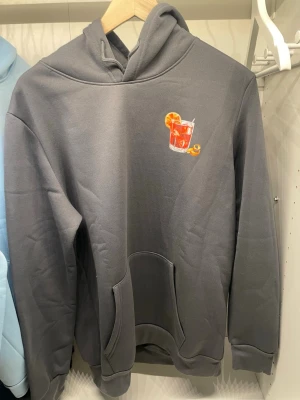 Grå hoodie med Negroni-tryck - Snygg grå hoodie med en färgglad Negroni-drink broderad på bröstet och stort tryck på ryggen med texten 'NEGRONI' och illustration av drinken. Hoodien har känguruficka och ribbade muddar. Perfekt för dig som gillar coola och unika prints.