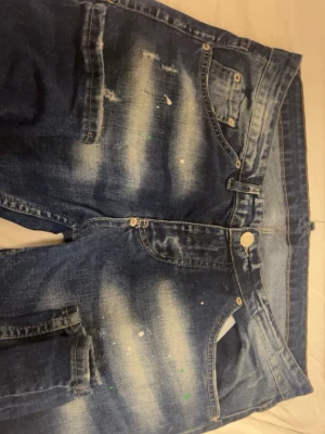 Blå slitna jeans från Dsquared2 - Säljer ett par blå jeans från Dsquared2 med slitningar och blekta partier. Jeansen har klassisk femficksdesign och feta färgstänk som ger en edgy look. Märkespatch i grönt med logga på bakfickan. Perfekt för dig som gillar streetstyle. Har aldirg använt bara en gång för o visa hur den ser ut när man har på sig den för o kunna visa en kund