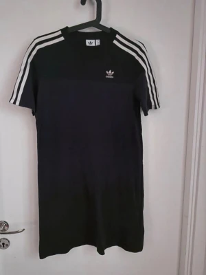 Svart t-shirtklänning Adidas - Svart t-shirtklänning från Adidas med klassiska vita ränder på axlarna och ärmarna. Adidas-logga på bröstet. Rak passform och korta ärmar. Tillverkad i mjukt material som känns skönt mot huden.
