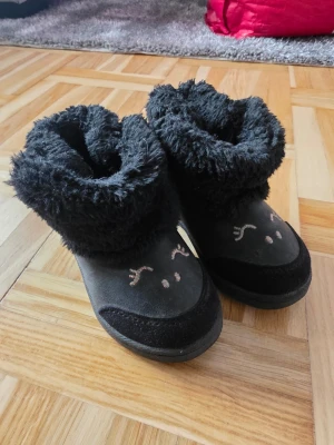 Svarta fluffiga boots med brodyr - Mysiga svarta boots med fluffigt foder och broderade detaljer framtill som liknar ett ansikte. Bootsens utsida är i mjukt material och de har en hög, bred krage i fuskpäls. Perfekta för att hålla fötterna varma under kalla dagar.