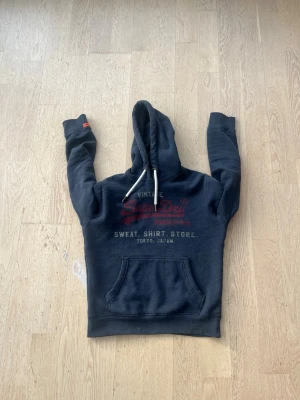 Mörkblå Superdry hoodie med tryck - Mörkblå hoodie från Superdry med stor röd och vit logga på bröstet samt japansk text. Klassisk känguruficka framtill och vita dragsnören i huvan. Mjuk insida och ribbade muddar vid ärmar och nederkant. Perfekt för en avslappnad streetwear-look.
