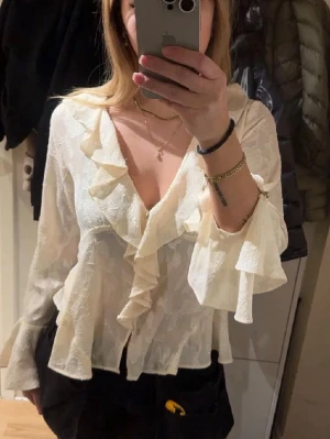 Vit volangblus från Zara - Säljer en vit blus med djup v-ringning och volangdetaljer längs halsringningen och ärmsluten. Blusen har ett lätt transparent tyg med subtilt mönster och långa ärmar.