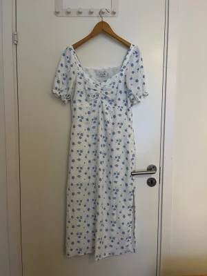 Vit blommig långklänning med puffärm - Vit långklänning med blått blommönster och puffärmar. Klänningen har v-ringning och en slits. Perfekt för sommaren🌸🌸 Klänningen är från Na-kd i storleken M, mycket fint skick✨