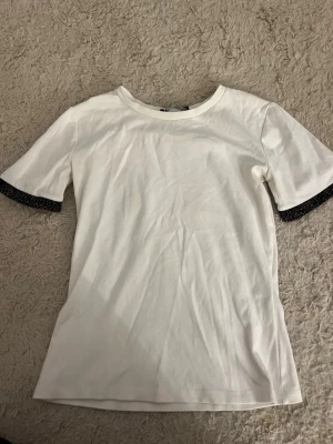 Vit t-shirt med glittriga ärmslut - Snygg vit t-shirt med korta ärmar och glittriga svarta detaljer vid ärmsluten.  Tröjan är i bra skick men har en lite fläck på baksidan av tröjan, syns på sista bilden.