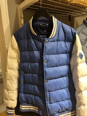 Blå och vit pufferjacka från GANT - Snygg pufferjacka från GANT i blått med vita ärmar och ribbade muddar. Jackan har tryckknappar framtill, collegekrage och GANT-logga på ena ärmen. Perfekt för kalla vinterdagar och ger en sportig look. Obs! Endast använd 2-3 gånger. Inköpt för drygt 2000kr för ett år sedan.