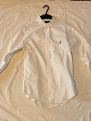 Vit skjorta från Ralph Lauren - Klassisk vit skjorta från Ralph Lauren med button-down krage och broderad logga på bröstet. Skjortan har lång ärm, knappar framtill och är tillverkad i mjuk bomull. Perfekt till både jeans och chinos för en clean look.