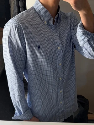 Blårutig skjorta från Ralph Lauren - Klassisk blårutig skjorta från Ralph Lauren med button-down krage och broderad logga på bröstet. Skjortan har långa ärmar, vita knappar och är tillverkad i mjuk bomull. Perfekt för en clean och stilren look.