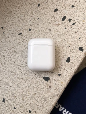 Apple AirPods med laddningsetui - Säljer ett par Apple AirPods med det klassiska vita laddningsetuiet. Etuiet är kompakt, har rundade kanter och en smidig knapp på baksidan för enkel parkoppling. Perfekt för dig som vill ha trådlösa hörlurar med stilren design och bra ljud.