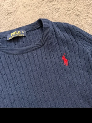 Marinblå kabelstickad tröja Polo Ralph Lauren - Kabelstickad marinblå tröja från Polo Ralph Lauren med klassisk röd logga broderad på bröstet. Tröjan har rund halsringning, långa ärmar och ribbade muddar. Perfekt för dig som gillar stilrena och tidlösa plagg.