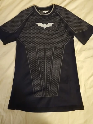 Batman Dark Knight cosplay t-shirt - Svart funktions-tshirt från Youngla med Batman Dark Knight Trilogy-tema. Tröjan har korta ärmar, grått mönster och Batman-logga på bröstet. Tillverkad i stretchigt och snabbtorkande material, perfekt för träning eller sport. Med denna kommer du vara kungen på gymmet! Den är äkta kan visa kvitto ifall det behövs anvönde den en gång och insåg att det var inte min grej med compression!