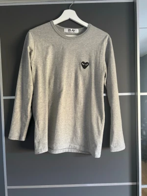 Grå långärmad tröja Comme des Garçons - Snygg grå långärmad tröja från Comme des Garçons PLAY med det ikoniska svarta hjärtat med ögon broderat på bröstet. Tröjan har rund halsringning och är tillverkad i mjukt bomullsmaterial. Perfekt för en clean och stilren look.
