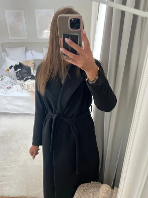 Svart lång kappa med knytskärp - Super snygg kappa från bikbok! 25% ull. Nopprat därav bra pris!💕💕 köpt på bikbok för 1000
