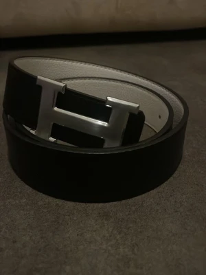 Svart skärp med H-spänne - Snyggt svart bälte med stort H-format metallspänne. Bältet har en stilren design och insidan är ljusare, vilket ger en cool kontrast. Perfekt accessoar för att lyfta din outfit. Du kan byta sida så den också blir vit!!!