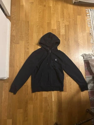 Blå hoodie från Polo Ralph Lauren - Säljer en blå hoodie från Polo Ralph Lauren med dragkedja framtill och klassisk huva. Tröjan har det ikoniska lilla vita Polo-logotrycket på bröstet och två fickor fram. Perfekt för en avslappnad och stilren look.
