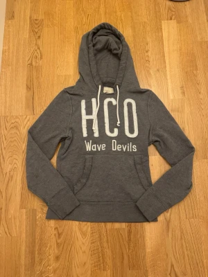 Grå Hollister hoodie HCO Wave Devils - En riktigt fett hollister hoodie i en mörk grå ton. Den är väldigt eftertraktad och knappt använd. Den är i Storlek S. Hör av vid frågor eller funderingar, kan även tänka mig diskutera pris