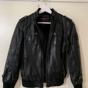 Svart skinnjacka från Katakit - Säljer en svart skinnjacka från Katakit med dragkedja och flera fickor framtill. Jackan har långa ärmar, ribbade muddar och dekorativa knappar på axlarna. Perfekt för dig som vill ha en cool och stilren look.