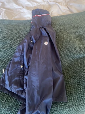 Marinblå vindjacka från Moncler, aldrig använd - Snygg marinblå vindjacka från Moncler med huva och vita samt röda detaljer på huvan. Jackan har dragkedja, tryckknappar och elastiska muddar vid ärmsluten. Klassisk Moncler-logga på ärmen och praktiska fickor framtill. Säljer eftersom att storleken inte passade för mig 