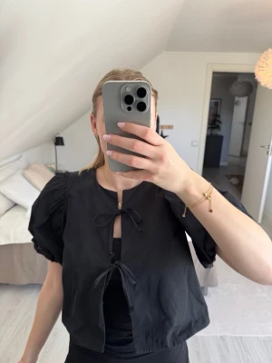 Svart puffärmad blus med knyt - Trendig svart blus med puffiga korta ärmar och två knytband framtill. Blusen har en lös passform och är tillverkad i ett lätt tyg som ger en luftig känsla. 