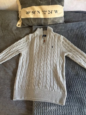 Ljusgrå kabelstickad tröja Polo Ralph Lauren - Ljusgrå kabelstickad tröja från Polo Ralph Lauren med halv dragkedja och hög krage. Tröjan har ett klassiskt flätmönster och den ikoniska blå logotypen broderad på bröstet. Ribbstickade muddar vid ärmslut och nederkant. Perfekt för sommar kvällar