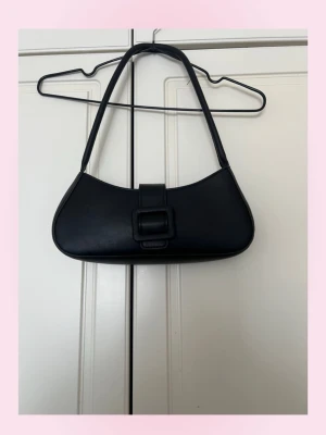 Svart axelremsväska i skinnimitation - 💕Stilren svart axelremsväska i skinnimitation med avlång form och bred axelrem. Väskan har ett lock med dekorativt spänne framtill och minimalistisk design utan synliga loggor. Perfekt för dig som gillar en clean och trendig look.💗