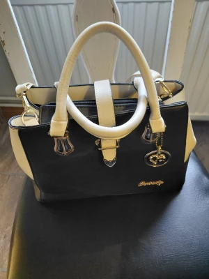 Nästan ny Svart och vit handväska Darbaoly - Nästan ny Snygg handväska från Darbaoly i svart och vitt konstläder. Väskan har två rejäla handtag, guldfärgade metalldetaljer och en dekorativ berlock. Klassisk fyrkantig form och dragkedja upptill. Perfekt för dig som gillar stilrena accessoarer.