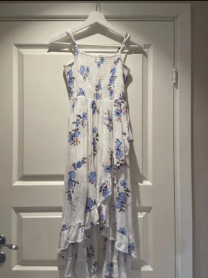 Vit blommig långklänning med volang - Romantisk långklänning i vitt med blått blommönster och smala axelband. Klänningen har smockad överdel, volangkant nertill och är tillverkad i ett lätt material som passar perfekt för sommaren.