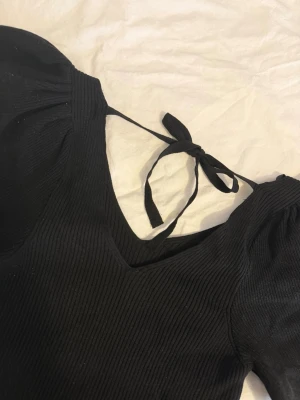 Svart ribbad topp med puffärm och knyt - Trendig svart ribbad topp med puffärmar och v-ringning. Toppen har snygg knytning i nacken och är tillverkad i ett stretchigt material som sitter snyggt på kroppen. Perfekt för dig som gillar en stilren men ändå unik look.